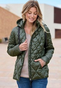 Grünes gesteppte Jacke mit Kapuze, ausgestattet mit einem Reißverschluss vorne, Seitentaschen und einer glatten Textur. Getragen über einem hellbeigen Oberteil und blauen Jeans.