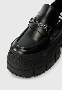 Mocassins pretos de plataforma com um acabamento brilhante, apresentando uma tira larga e acabamentos metálicos. Sola de borracha texturizada com a inscrição "BUFFALO".