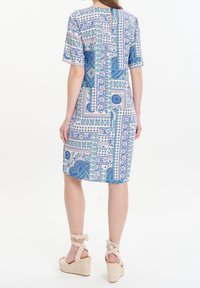 Frau in einem knielangen, kurzärmeligen Kleid mit blau-grünem Patchwork-Muster, kombiniert mit beigen Keilsandalen mit Knöchelriemchen.