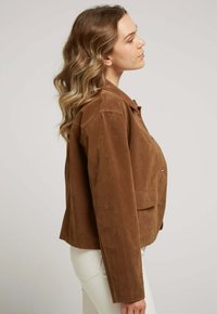 Veste en daim marron avec col, fermeture à boutons sur le devant et deux poches latérales. Ajustement cintré avec une texture lisse et des coutures plates.