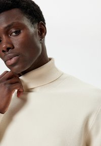 Sweter typu golf w kolorze kremowym z prążkowanym kołnierzem i gładką fakturą, noszony przez osobę z krótkimi, ciemnymi włosami i kolczykiem.