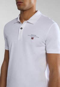 Hvid polo T-shirt med korte ærmer, blødt bomuldsstof, med knaplukning, broderet logo og et lille flagdetalje på brystet.
