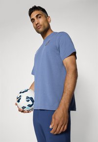 Blå bomulds-T-shirt med kortærmer, logodetalje og afslappet pasform; parret med blå træningsbukser og holder en mønstret fodbold.