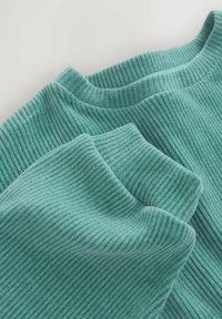 Primo piano di un tessuto a coste di un colore teal che mostra i dettagli del colletto e dei polsini delle maniche di un indumento a maniche lunghe.
