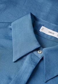 Chemise bleu clair avec un col et un seul bouton fermé au niveau du cou ainsi qu'une étiquette de marque visible à l'intérieur du col.