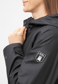 Zwarte waterdichte jas met een capuchon, voorzien van een ritssluiting en drukknoopsluiting. Opvallende logo patch op de mouw met een architectonisch ontwerp.