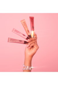 Five tubes of matte blush in varying shades of pink and coral, held by a hand with a crystal bracelet, against a pink background.