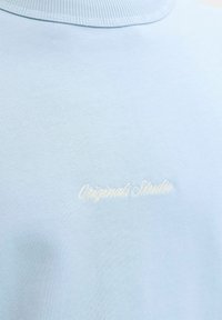 Šviesiai mėlynas medvilninis marškinėlis su siauru apykakle. Krūtinėje siuvinėtas baltas „Originals Studio“ logotipas.