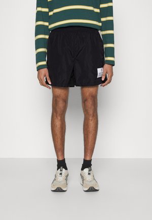 Muž oblečený v černých sportovních šortkách s bílou logovou nášivkou, zelenožlutém pruhovaném tričku s dlouhým rukávem, černých ponožkách a béžových teniskách.