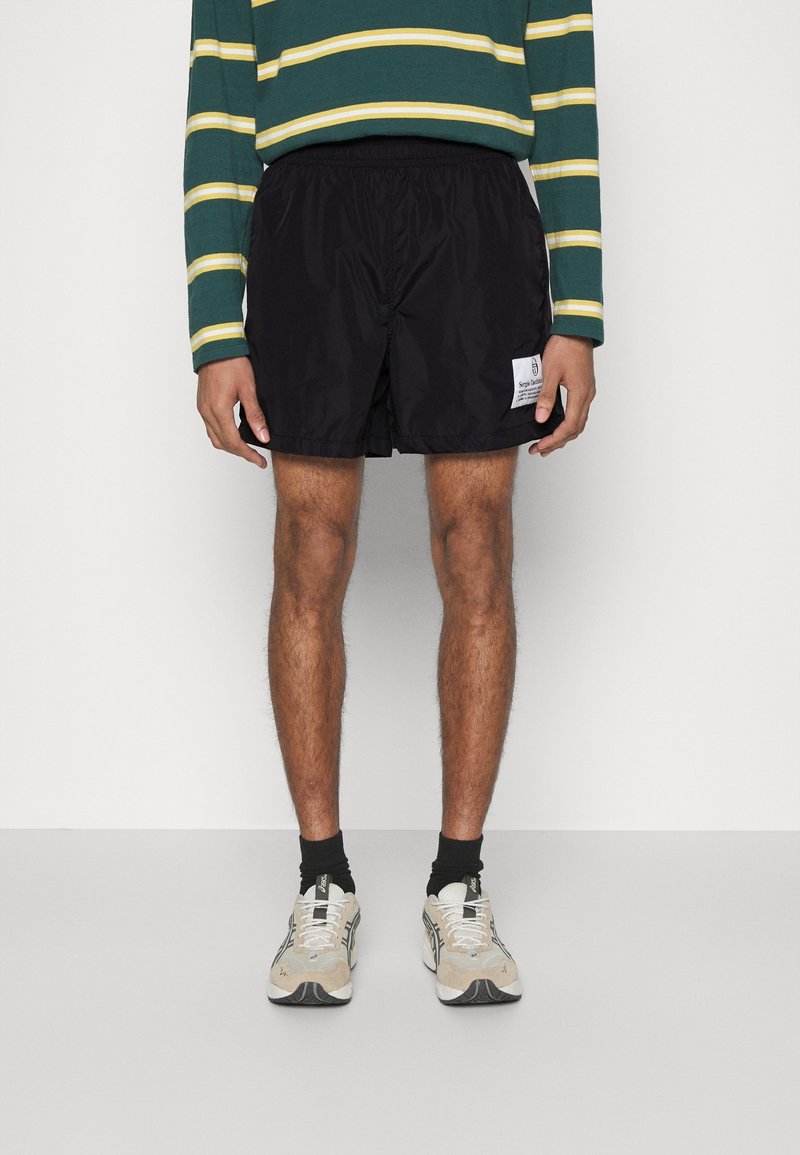 Homme portant un short de sport noir avec un patch logo blanc, un t-shirt à manches longues rayé vert et jaune, des chaussettes noires et des baskets beiges.