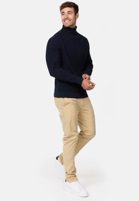 Granatowy sweter z dzianiny typu golf z teksturowanym wzorem, zestawiony z jasnobeżowymi chinosami i białymi trampkami. Model stoi w zrelaksowanej pozie.