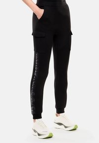 Pantaloni da jogging neri in materiale morbido con tasche laterali, caratterizzati da un logo in rilievo verticale sulla gamba sinistra e caviglie a costine.