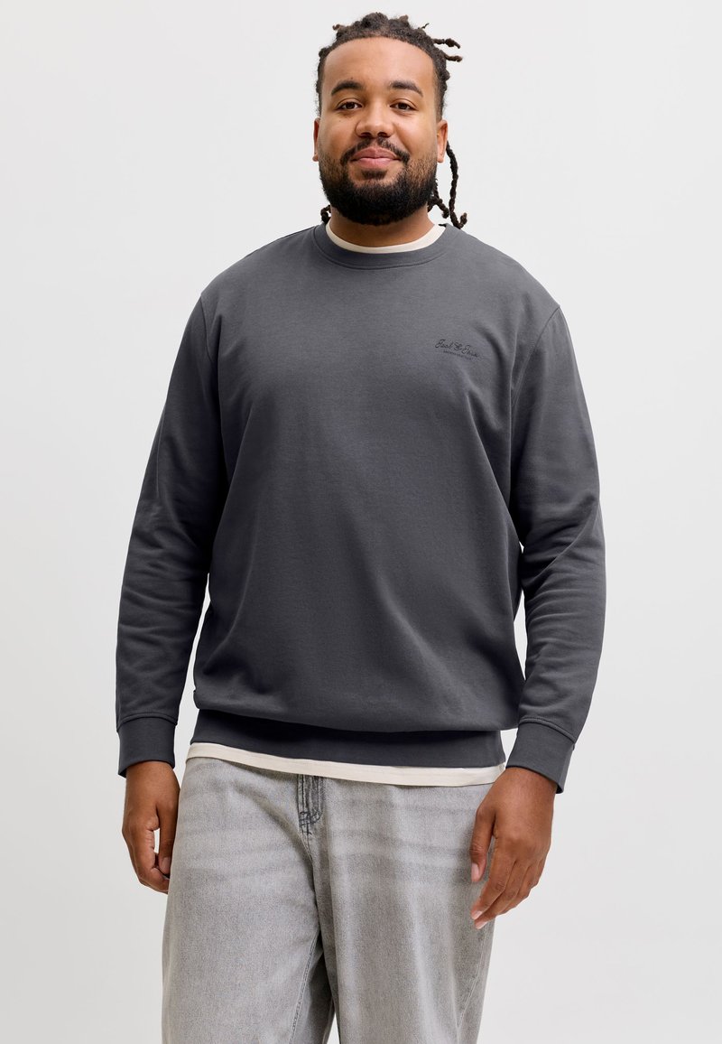 Homme aux cheveux tressés et à la barbe, portant un sweat-shirt gris foncé à col rond et un jean gris clair, debout devant un fond clair uni.