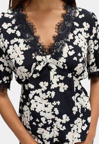 Blusa floreale a maniche corte con tessuto nero decorato da fiori bianchi e dettagli rosa. Bordo in pizzo lungo il collo e le maniche. Dettaglio a fiocco al centro.