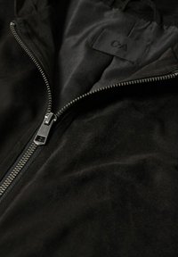 Gros plan d'une veste en daim noire partiellement dézippée avec une fermeture éclair argentée et une doublure intérieure noire avec une étiquette de marque.