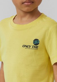 Geel katoenen t-shirt met een ronde afbeelding op de linkerborst met de tekst "ENKEL DE" en "WILD DUNE WINDS WEET" in zwart en teal.