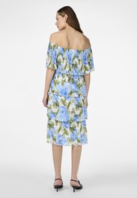 Robe à épaules dénudées, à volants, en tissu blanc, ornée de grands motifs floraux bleus avec des feuilles vertes. Texture légère et plissée.