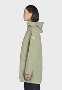 Veste imperméable vert clair avec capuche, poches latérales et un patch de marque sur la manche. Texture lisse et coupe décontractée.