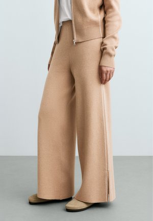 Femme portant un pantalon en maille beige à jambes larges avec des bandes blanches sur les côtés et des chaussures beiges, debout sur un sol gris contre un mur clair.