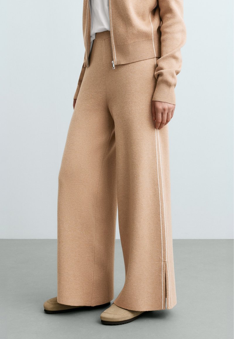 Femme portant un pantalon en maille beige à jambes larges avec des bandes blanches sur les côtés et des chaussures beiges, debout sur un sol gris contre un mur clair.