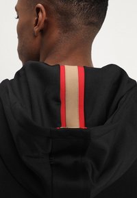 Detailní záběr na krk a hlavu osoby nosící černou mikinu s kapucí, na zadní straně kapuce je vertikální béžový a červený pruh.