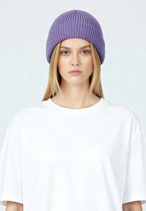 Jonge vrouw met lang blond haar, een paarse gebreide muts en een eenvoudig wit oversized T-shirt, die recht vooruit kijkt met een neutrale uitdrukking.