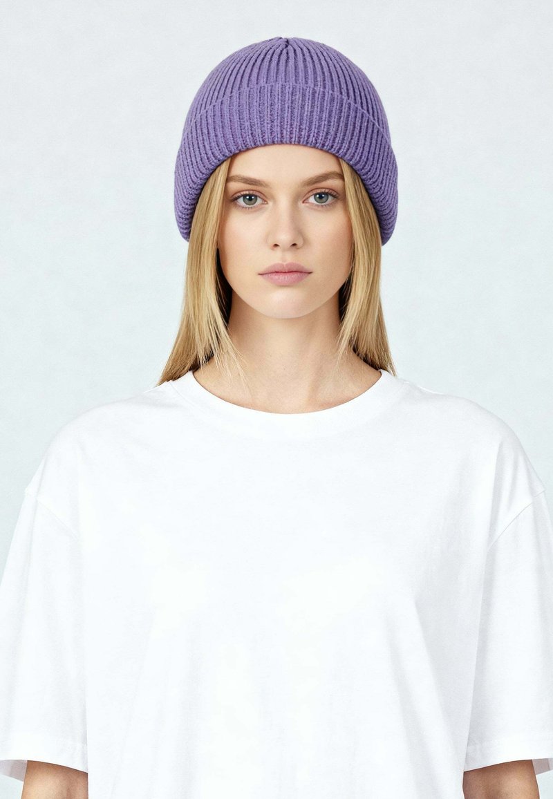 Giovane donna con lunghi capelli biondi che indossa un berretto viola lavorato a maglia e una maglietta bianca oversize semplice, rivolta in avanti con un'espressione neutra.