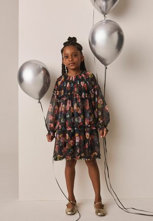Jong meisje in een donker doorschijnend bloemenjurkje met drie zilveren ballonnen tegen een effen lichte achtergrond, met gouden schoenen.