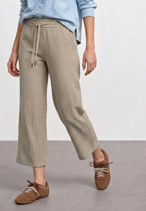 Persoon draagt beige getextureerde cropped broek met trekkoord en bruine veterschoenen, staand tegen een effen lichte achtergrond.