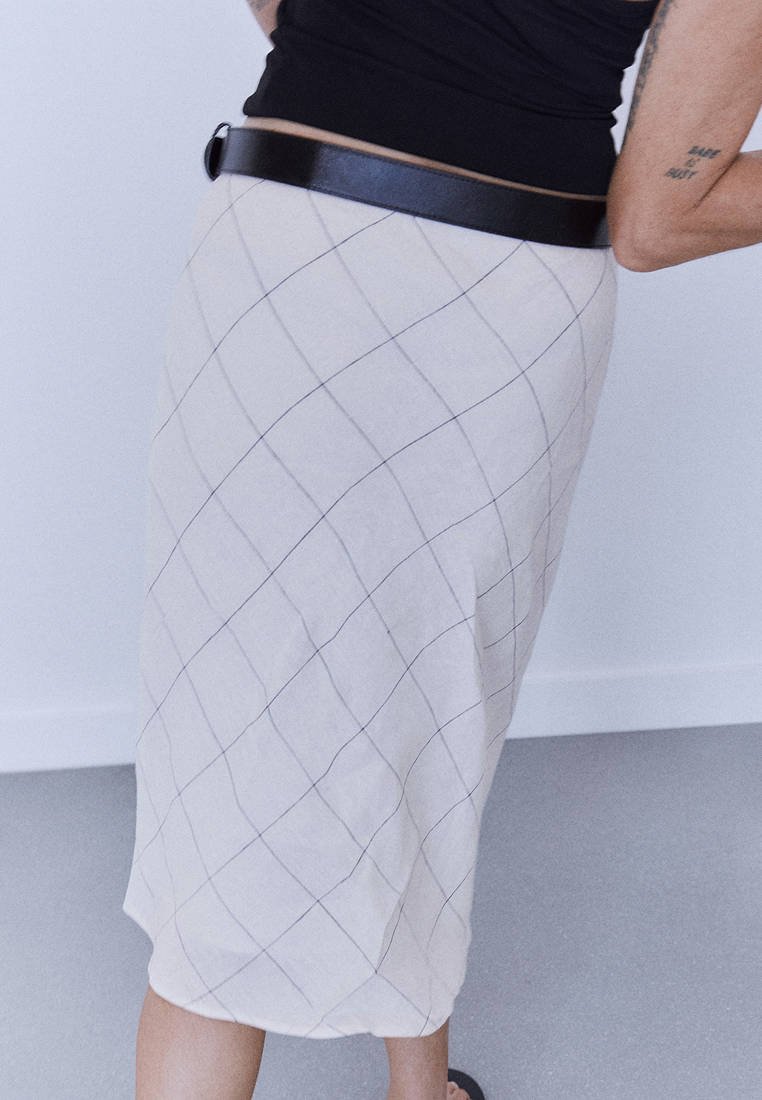Personne portant une jupe blanche à motif quadrillé noir, une ceinture noire et un haut noir, debout devant un fond clair uni.
