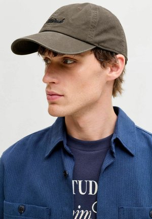 Brązowa bawełniana czapka z daszkiem z haftowanym napisem. Nosi się ją z niebieską, teksturowaną koszulą na guziki oraz ciemnym t-shirtem pod spodem.