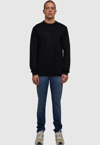 Schwarzes Langarm-T-Shirt mit Rundhalsausschnitt, kombiniert mit blauen Slim-Fit-Jeans und beigen Sneakers, vor einem schlichten grauen Hintergrund stehend.