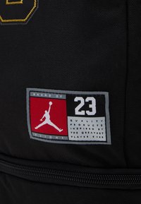 La etiqueta de tela negra presenta gráficos en blanco y rojo, que incluyen una silueta, el texto "23" y "Marca de Vuelo" en una superficie texturizada.