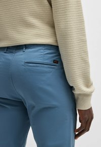 Light beige knitted sweater and blue chino pants with a back pocket featuring a red and black logo tag. Smooth texture, tailored fit.