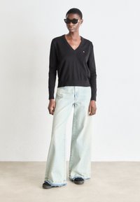 Pulover negru cu decolteu în V, asortat cu blugi largi de culoare albastru deschis. Blugii au o margine brută și o textură decolorată. Ochelarii de soare negri completează ținuta.