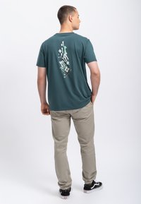 Teal crew-neck t-shirt met een grafisch ontwerp in het wit en groen op de achterkant. Gecombineerd met beige broek en zwarte sneakers.