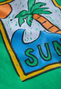 Grüner Stoff zeigt ein farbenfrohes grafisches Design mit einer Sonne, einer Palme und Wellen. Auffällige Texturen sind erhabene Konturen und lebendige Farben.