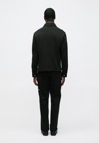 Mustast kraega jakk pikkade varrukatega, kantud tumedate teksade üle, vaadeldud tagantpoolt neutraalse tausta ees.