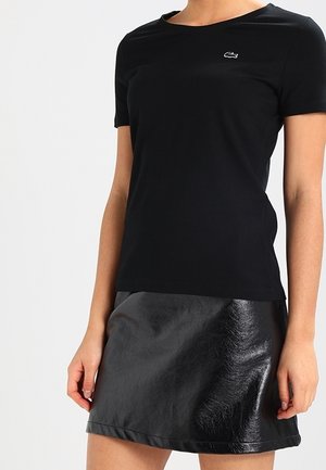 Nainen, jolla on yksinkertainen musta lyhythihainen paita ja kiiltävä musta nahkahame, seisoo yksinkertaisen vaalean taustan edessä.