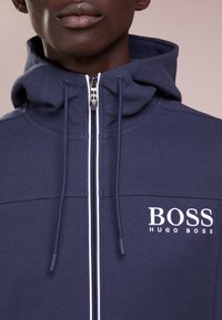 Marinblå hoodie med texturerad övre panel, vit dragkedja och justerbara snörningar. Har broderad "BOSS HUGO BOSS" på bröstet.