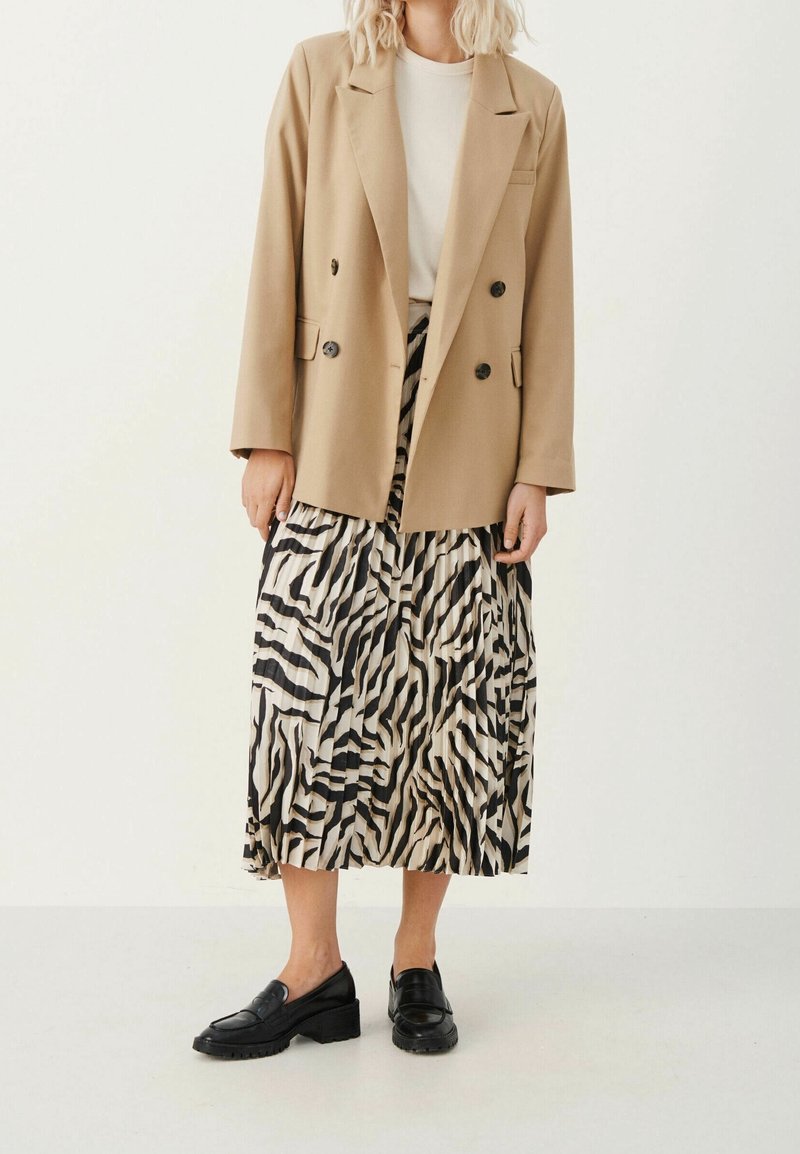 Personne portant un blazer beige, un haut blanc, une jupe midi à motif zébré noir et blanc et des mocassins noirs, debout devant un fond uni.