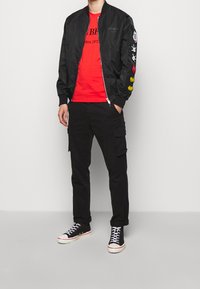 Chaqueta bomber negra con diseños bordados en las mangas, camiseta gráfica roja, pantalones cargo negros y zapatillas deportivas negras de caña alta con acentos blancos.