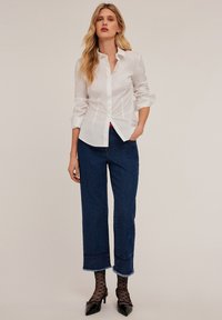 Camisa blanca de botones con mangas remangadas, combinada con unos vaqueros recortados de talle alto en azul oscuro con dobladillos deshilachados y costuras laterales. Zapatos negros de tacón.
