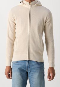 Beige zip-up hoodie with ribbed texture, fitted design, and two side pockets; paired with light blue denim jeans.