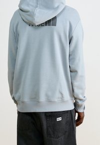 Felpa con cappuccio in pile azzurro chiaro con tasca a marsupio, con un logo nero sul retro. Polsini e orlo a costine, abbinata a jeans neri.