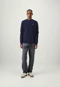 Sweatshirt azul marinho de manga longa com decote redondo e um logótipo amarelo. Combinado com jeans cinzentos e ténis brancos com detalhes em beje.