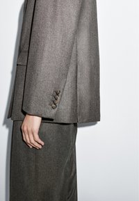 Hnědý blejzr z vlněné směsi s rovnými liniemi, který má na manžetách čtyři černé knoflíky a jemnou strukturu. K obleku patří také kalhoty.