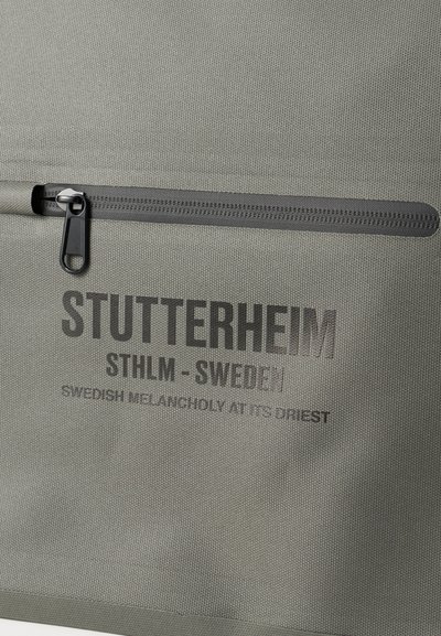 Pelēks auduma soma ar rāvējslēdzēja kabatu, kurā ir izspiests teksts: "STUTTERHEIM STHLM - Zviedrija" un "ZVIEDRU MELANHOLIJA VISĀ PILNĪBĀ."