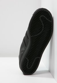 Černá podrážka tenisky s texturovaným vzorem herringbone. Vyrobena z gumy, má vyvýšené drážky a na boku je ražené logo Adidas.