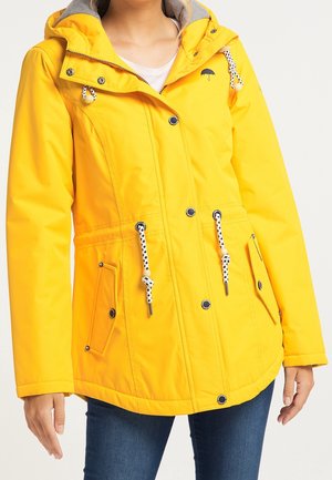 Femme portant une veste de pluie jaune vif avec des boutons, une taille ajustable par cordon, des poches à rabats et un petit logo parapluie noir sur la poitrine.